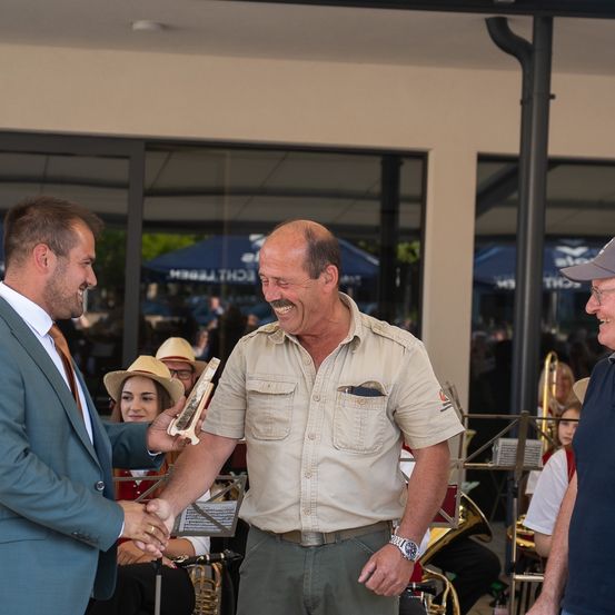Zwei Männer in Anzügen schütteln sich die Hände, einer hält eine Trophäe, stehen vor einem Veranstaltungsort mit einer Band und Publikum im Hintergrund.