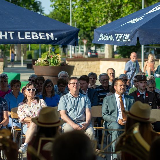 Eine Outdoor-Veranstaltung mit vielen Menschen, die unter blauen Schirmen sitzen. Einige tragen Hüte, Brillen und Anzüge. Der Mann im Anzug hält ein Mikrofon und spricht. Ein blauer Schirm mit weißer Beschriftung ist im Hintergrund.