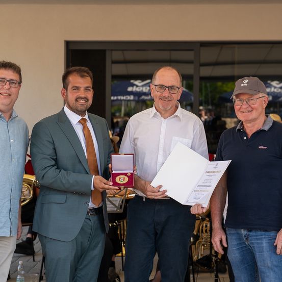 Vier Männer stehen zusammen und lächeln für ein Foto. Der Mann auf der linken Seite trägt ein blaues Hemd, der zweite einen Anzug mit Krawatte, der dritte ein weißes Hemd und der vierte ein marineblaues Polohemd mit einer Mütze. Sie halten ein Zertifikat und eine Medaille. Hinter ihnen sind Instrumente und Menschen durch eine Glastür sichtbar.