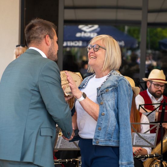 Ein Mann in einem Anzug überreicht einer lächelnden Frau, die eine Brille und eine Jeansjacke trägt, einen hölzernen Preis, während eine Band im Hintergrund spielt.