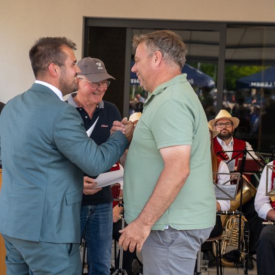 Zwei Männer schütteln sich die Hände und halten Essen vor einem Mann in einem Anzug, während ein Mann mit Hut hinter ihnen steht und Musiker im Hintergrund Instrumente spielen.
