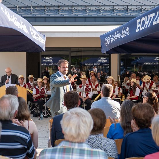 Ein Mann spricht an einem Rednerpult, während eine Brassband spielt. Das Publikum sitzt in Stühlen unter blauen Schirmen. Die Szene findet im Freien vor einem Gebäude statt.