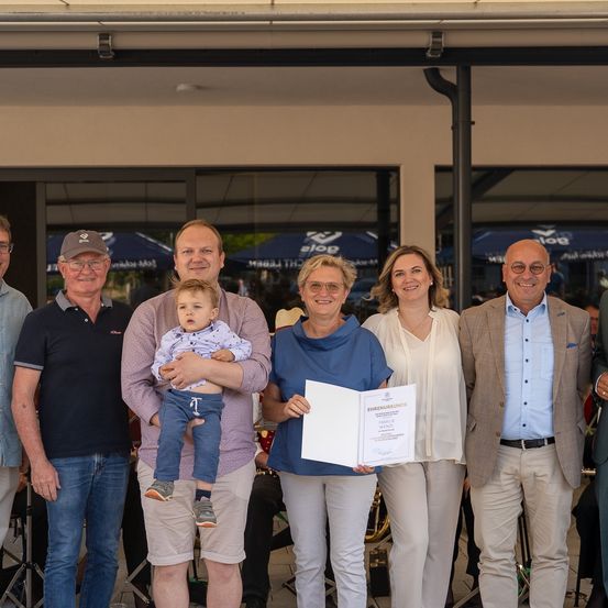 Eine Gruppe von Erwachsenen und einem Kind stehen lächelnd vor einem Gebäude. Eine Frau hält eine Urkunde. Die Erwachsenen tragen formelle Kleidung. Dahinter ist ein verschwommener Blick auf ein weiteres Gebäude und einen Baum.
