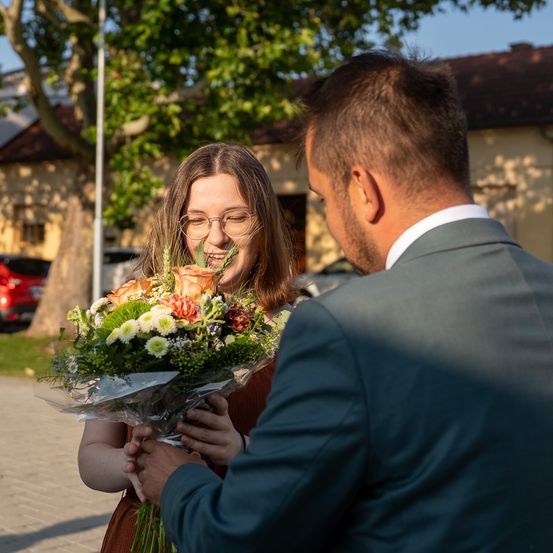 Ein Mann in einem Anzug überreicht einer lächelnden Frau mit Brille einen Blumenstrauß. Sie stehen vor einem Haus mit geparkten Autos.