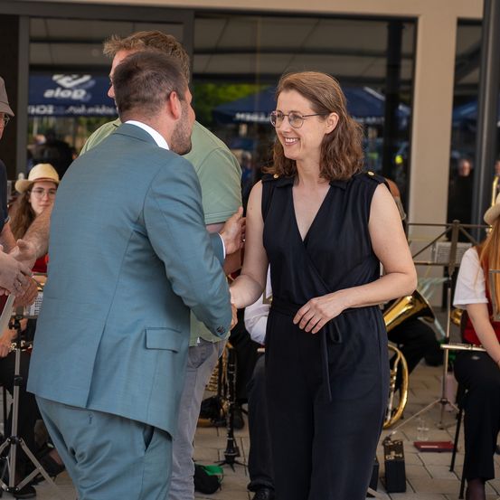 Ein Mann in einem blauen Anzug gibt einer Frau in einem schwarzen Kleid die Hand vor einer Band. Hinter ihnen sitzt eine Frau in einem Hut, und es sind weitere Personen im Hintergrund zu sehen. Musikinstrumente sind sichtbar.