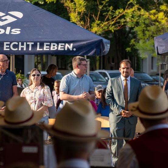 Eine Gruppe von Menschen unter einem Zelt, möglicherweise bei einer Veranstaltung. Ein Mann in Anzug steht mit gefalteten Händen. Eine Frau und zwei Männer stehen in der Nähe. Autos und Bäume sind im Hintergrund.