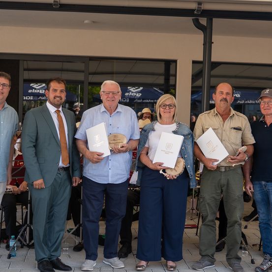 Eine Gruppe von sechs Personen in formeller Kleidung posiert für ein Foto vor einem Gebäude und hält Urkunden in den Händen. Dahinter sitzen verschwommene Figuren in der Nähe eines Zelts, während ein Mann bei einem Mikrofon steht.