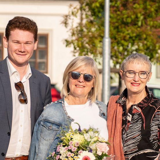 Ein junger Mann und zwei Frauen stehen lächelnd zusammen. Die Frau auf der linken Seite hält einen Blumenstrauß. Sie stehen draußen vor einem Gebäude und Bäumen.