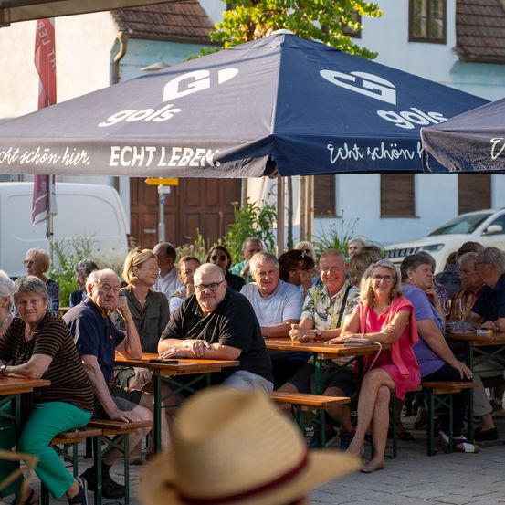 Eine Gruppe älterer Menschen sitzt an Tischen unter blauen Sonnenschirmen in einem Außenbereich. Die Sonnenschirme haben weiße Beschriftungen. Hinter ihnen sind zwei weiße Autos geparkt.
