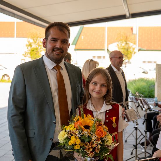 Ein Mann und ein junges Mädchen mit Blumen lächeln für ein Foto. Hinter ihnen beobachtet ein Mann mit Brille, und Musiker spielen Instrumente auf einer Bühne.