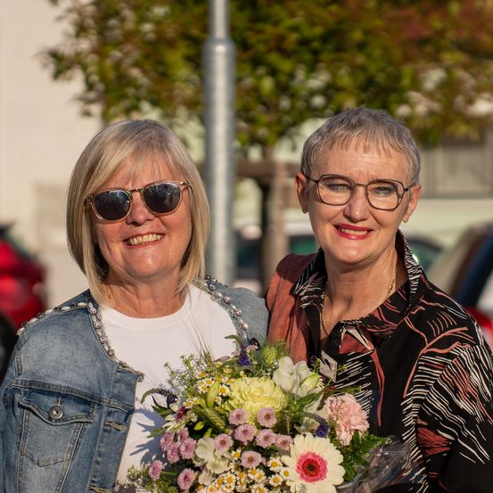 Zwei Frauen stehen eng beieinander, lächeln und halten einen Blumenstrauß. Die Frau auf der linken Seite trägt eine Jeansjacke und eine Sonnenbrille, während die andere Frau ein gemustertes Hemd und eine Brille trägt. Sie sind draußen in der Nähe eines Baumes.