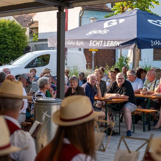 Eine Gruppe von Menschen sitzt unter einem blauen Schirm, vermutlich in einer Biergarten, und eine Frau steht in der Nähe mit einem Notenständer.