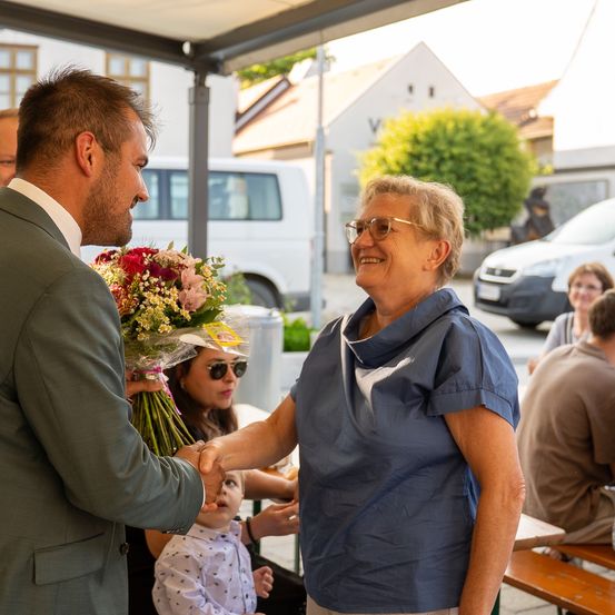 Ein Mann in einem Anzug, der Blumen hält, schüttelt die Hand einer lächelnden Frau mit Brille in einer Außenumgebung. Hinter ihnen sitzt ein kleines Kind und zwei Personen sitzen an einem Tisch. Im Hintergrund steht ein Lieferwagen und ein Haus.