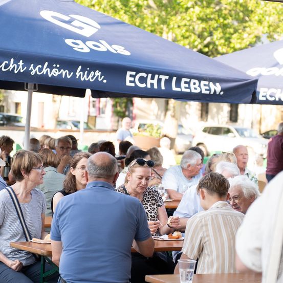 Eine Gruppe von Menschen, darunter ältere Erwachsene, sitzt unter blauen Sonnenschirmen in einem Outdoor-Restaurant. Sie essen und unterhalten sich. Die Sonnenschirme haben weißen Text, der 'ECHT LEBEN' besagt. Im Hintergrund sind Bäume und geparkte Autos zu sehen.
