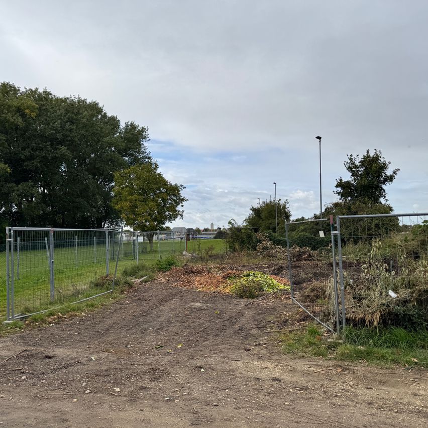 Ein leeres Grundstück mit einem Erdweg, umgeben von einem Metallzaun. Im Hintergrund sind Bäume und ein grünes Feld zu sehen. Ein Straßenlicht steht in der Nähe.