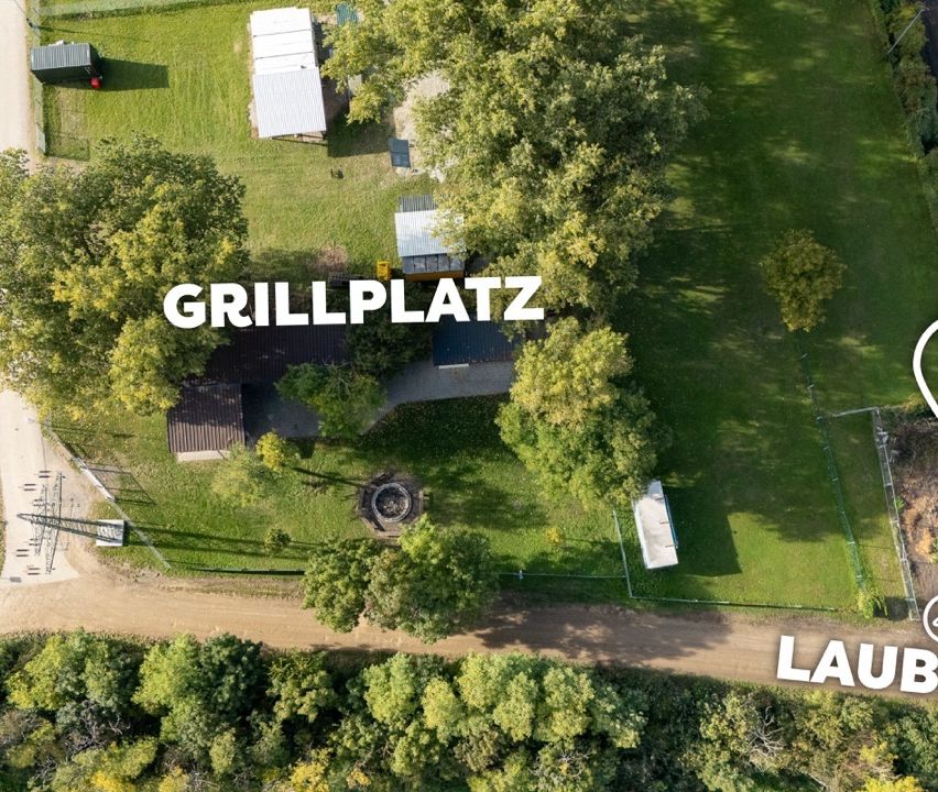 Eine Luftaufnahme eines Hinterhofs mit einem großen grünen Rasen, Bäumen und einem Feuerplatz. Die Worte 'GRILLPLATZ' sind prominent angebracht.