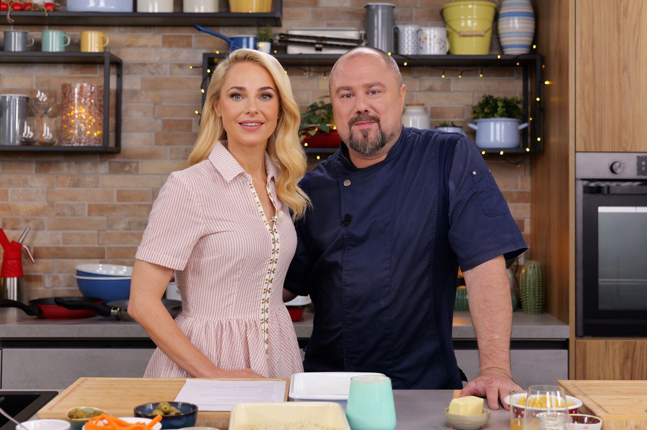 Ein Mann und eine Frau stehen lächelnd in einer Küche. Hinter ihnen befinden sich Regale mit Küchenutensilien. Auf dem Tresen sind verschiedene Zutaten und Utensilien angeordnet, darunter Karotten, Oliven und eine Tasse.