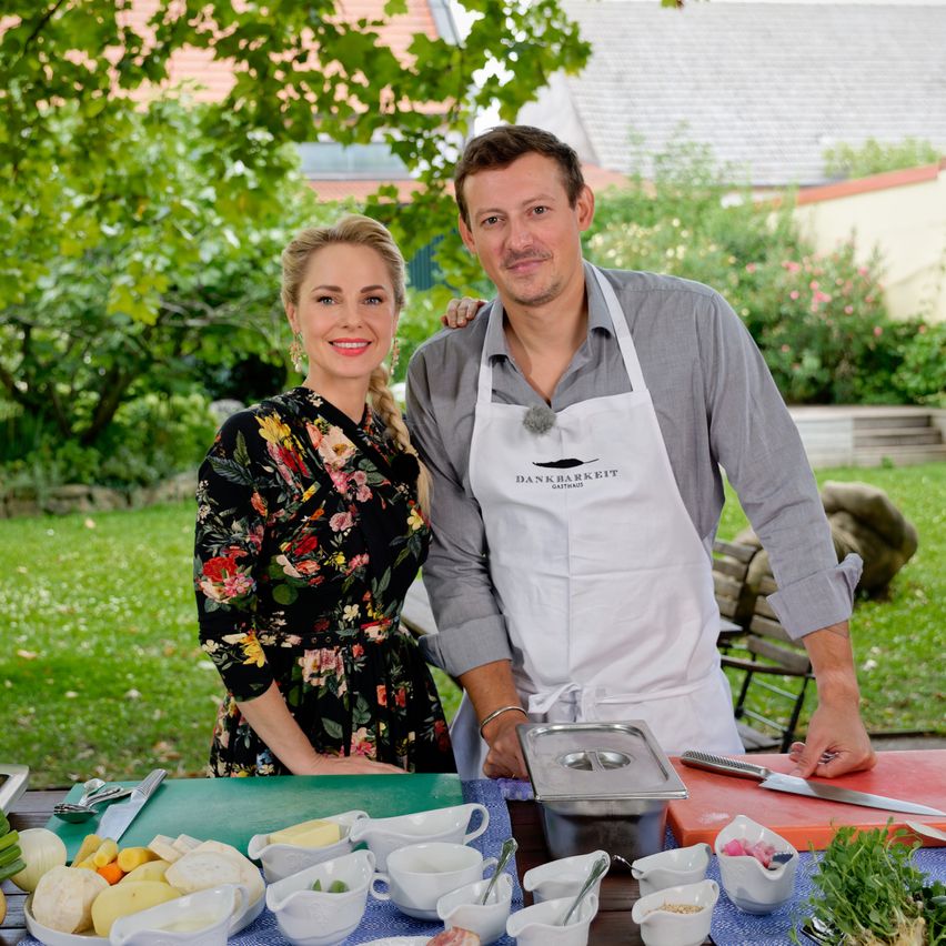 Ein Mann und eine Frau stehen in einem Garten und bereiten Essen auf einem Tisch mit verschiedenen Zutaten und Utensilien zu. Der Mann trägt eine Schürze mit dem Text DANKBARKEIT.