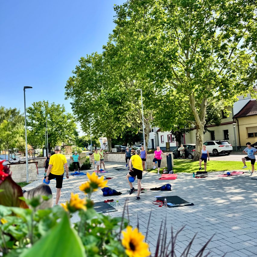 Eine Gruppe von Menschen macht ein Outdoor-Workout in einem Park mit Bäumen und Blumen. Sie machen Übungen mit Hanteln und Matten.