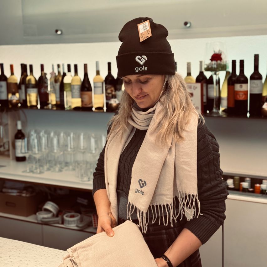 Eine Frau in einer schwarzen Mütze und einem Schal hält einen beigefarbenen Gegenstand vor einem Weinladen. Hinter ihr stehen Flaschen Wein in Regalen.