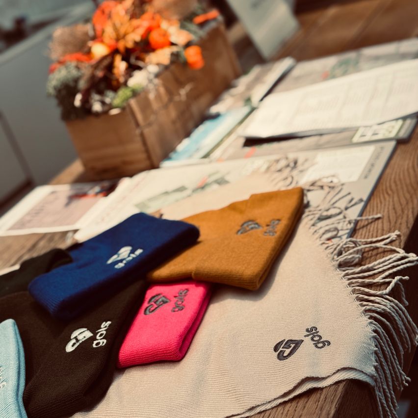 Ein Holztisch mit verschiedenen Gegenständen darauf, einschließlich eines Schal, mehrerer Mützen und verstreuten Papieren, mit einem Holzkasten im Hintergrund, der Blumen enthält.