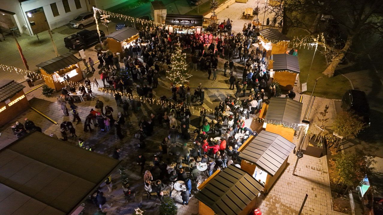 Eine Luftaufnahme eines überfüllten Weihnachtsmarktes mit zahlreichen Ständen und einem großen zentralen Weihnachtsbaum, umgeben von funkelnden Lichtern und festlichen Dekorationen.
