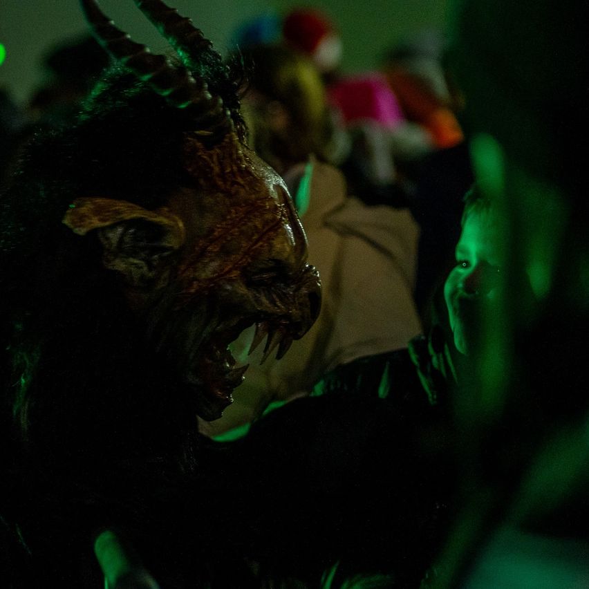 Eine Person mit einer gehörnten Maske und offenem Mund steht in einem schwach beleuchteten Raum nah an einem Kind.