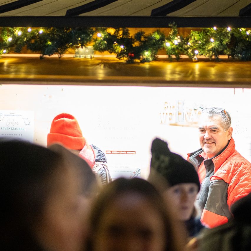 Ein Mann in einer orangefarbenen Jacke lächelt, während er mit Menschen in einer festlichen, überdachten Umgebung interagiert. Oben schmücken festliche Lichter und Pflanzen die Decke.