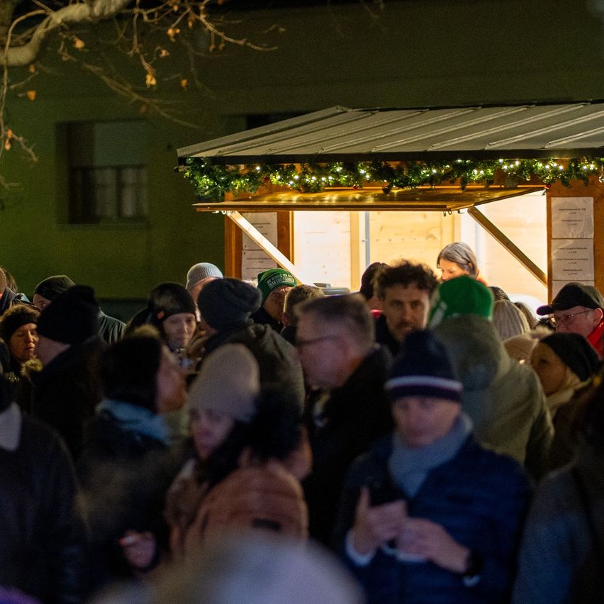 Eine Menschenmenge in Winterkleidung steht vor einem geschmückten Stand mit Lichtern und Grün. Der Stand befindet sich in der Nähe eines Baumes und eines Gebäudes.