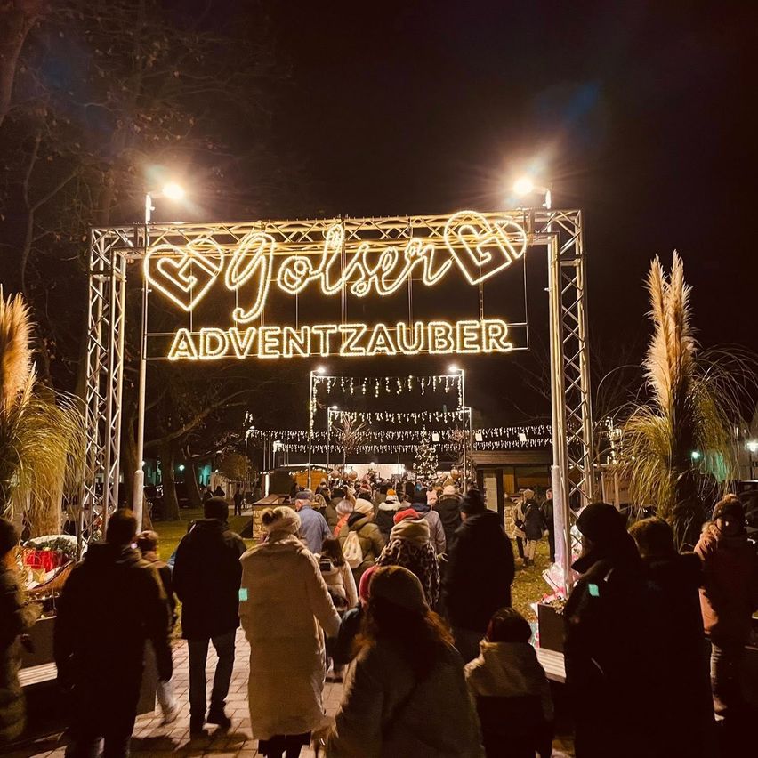 Beleuchtetes Schild mit 'Joliser Adventzauber' in Herzform. Menschen gehen nachts vor Ständen vorbei.