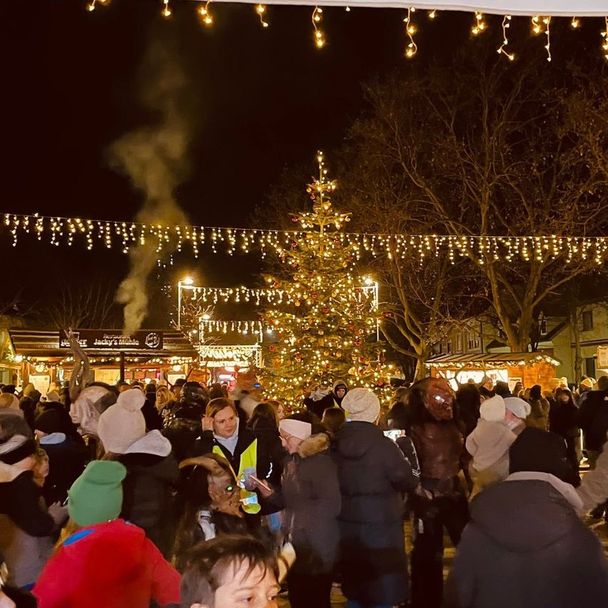 Eine Menschenmenge versammelt sich nachts um einen geschmückten Weihnachtsbaum. Der Baum ist mit Lichtern beleuchtet und Rauch steigt auf. Das Gebiet ist mit anderen festlichen Lichtern gefüllt.