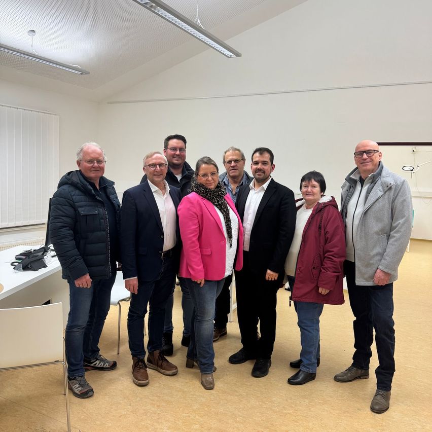 Eine Gruppe von sieben Personen, darunter Männer und Frauen, steht in einem Raum. Sie sind alle geschäftlich gekleidet.