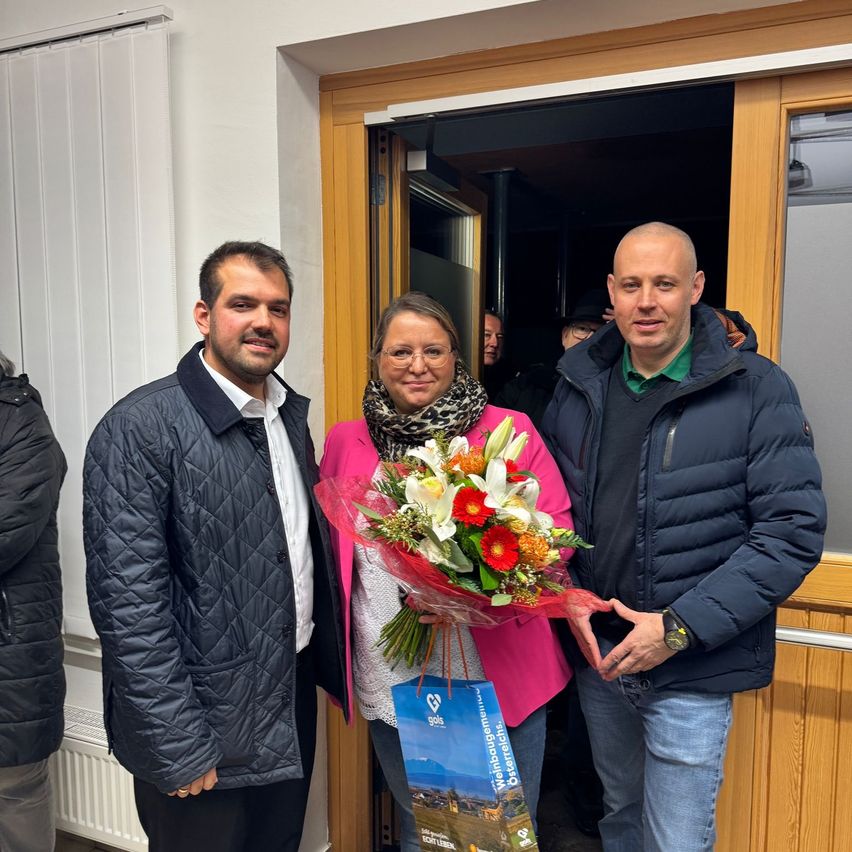 Drei Personen stehen lächelnd vor einer offenen Tür. Eine Frau in der Mitte hält einen Blumenstrauß. Hinter ihr hält ein Mann eine Tasche. Die Umgebung scheint ein Haus zu sein.