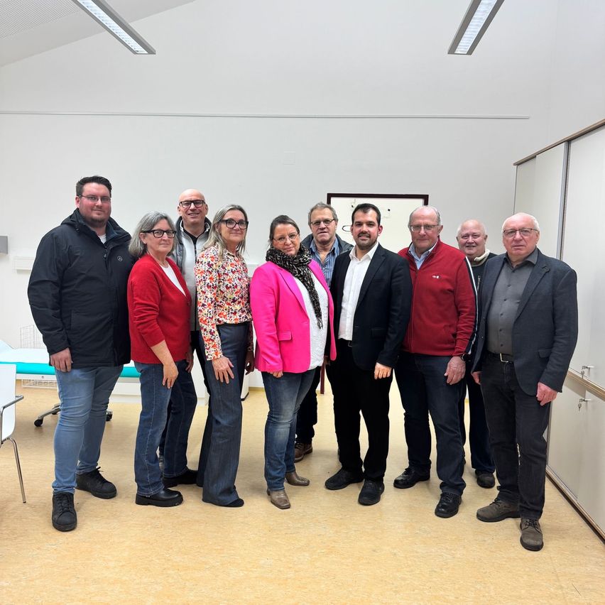 Eine Gruppe von Menschen steht in einem Raum zusammen. Einige lächeln und andere schauen in die Kamera. Der Raum hat weiße Wände und einen Holzboden.