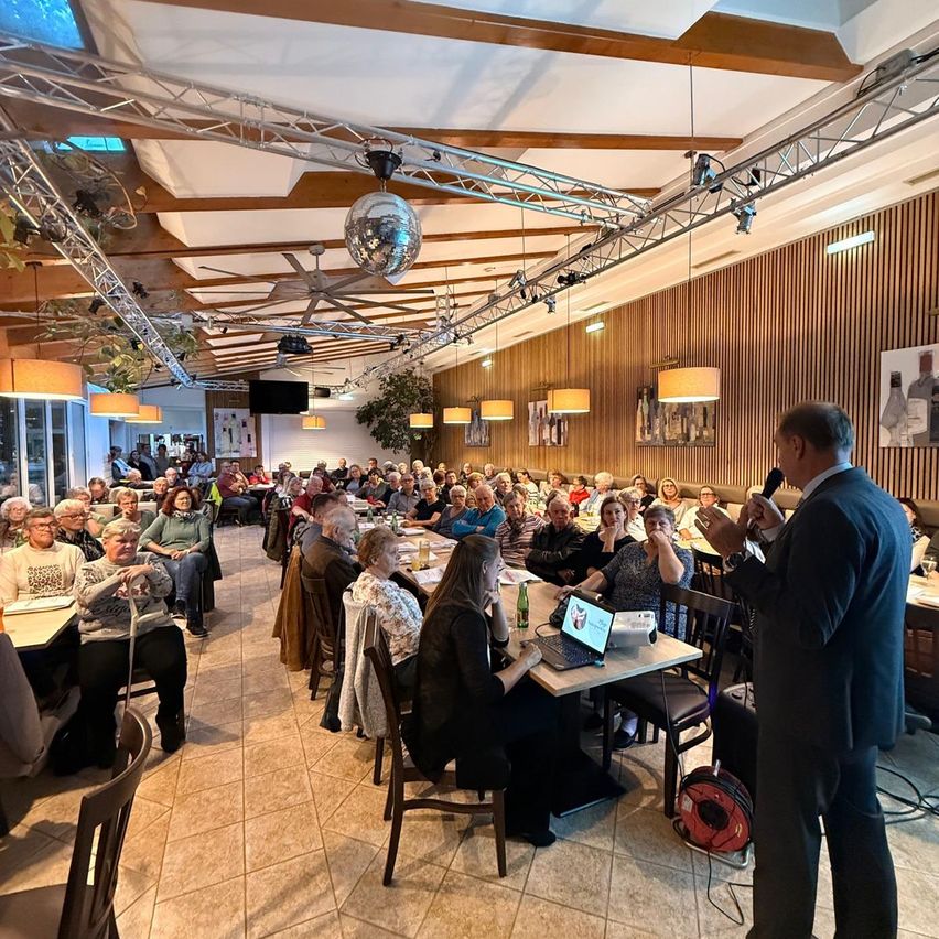 Ein Mann spricht in ein Mikrofon bei einer großen Versammlung in einem Raum mit Holzwänden, Pendelleuchten und einer Projektionsleinwand. Die Leute sitzen an Tischen und hören aufmerksam zu.