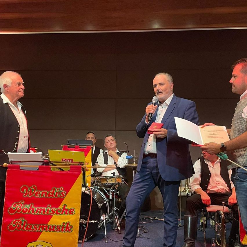 Eine Preisverleihung mit einer Band im Hintergrund. Ein Mann in einem blauen Anzug hält ein Mikrofon und ein Zertifikat, während andere in der Nähe stehen.