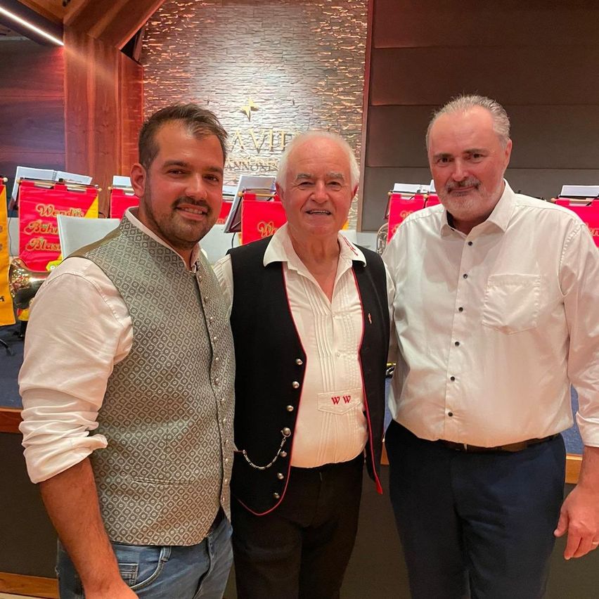 Drei Männer stehen zusammen in einem Raum mit einer Holzwand und lächeln für ein Foto. Der Mann auf der linken Seite trägt eine Weste und Jeans. Der Mann in der Mitte trägt ein weißes Hemd und eine schwarze Weste. Der Mann auf der rechten Seite trägt ein weißes Hemd und blaue Hosen.