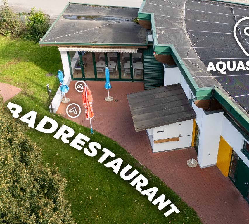 Ein Blick von oben auf das Aqua Restaurant mit Tischen und Stühlen draußen, grünen Dächern und Solarpanels auf dem Gebäude. Es gibt Sonnenschirme und einen Rasenbereich.