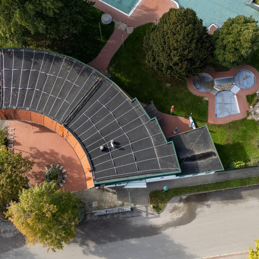 Eine Draufsicht auf einen großen Tennisplatz mit schwarzem Dach, umgeben von grünem Rasen und Bäumen. Einige Personen gehen an der Seite entlang.