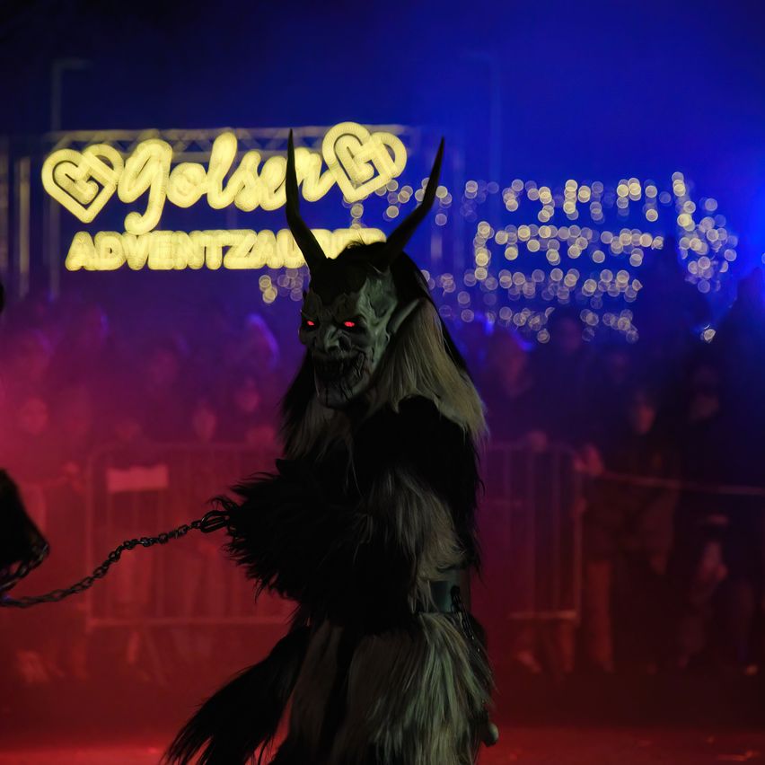 Eine Person in einem Kostüm mit Hörnern und einer Maske steht vor einer Menge mit blauem und rotem Hintergrund und einem Schild mit der Aufschrift 'Jolson Adventzaun'.