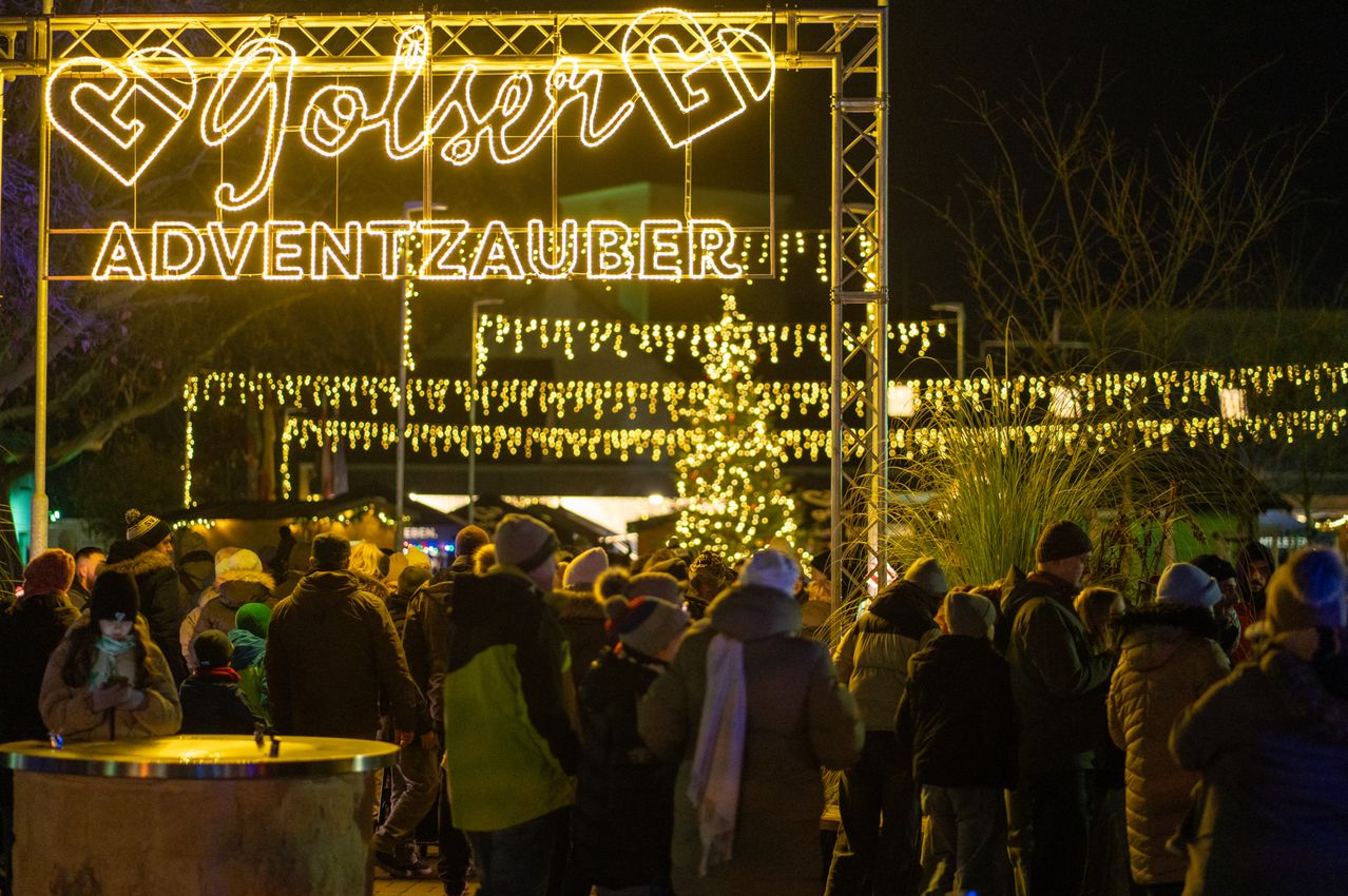 Eine Menschenmenge steht unter einem großen beleuchteten Torbogen mit 'Joulezauber' in Gelb. Es ist Nacht, und viele Weihnachtslichter erhellen den Bereich. Bäume sind mit Lichtern geschmückt, und es herrscht eine festliche Atmosphäre.
