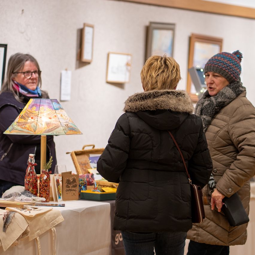 Drei Frauen, zwei in Wintermänteln, betrachten Gegenstände an einem Stand. Eine Frau hält eine Lampe. Der Stand hat einen Tisch mit Gegenständen und gerahmte Bilder an der Wand.