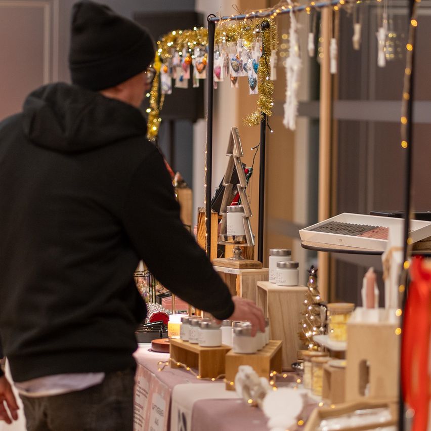 Ein Mann in einer schwarzen Kapuzenjacke steht an einem Tresen und betrachtet verschiedene Gegenstände. Der Tresen ist mit kleinen Gläsern, Ornamenten und einer Registrierkasse gefüllt. Der Raum ist mit Girlanden und Lichtern dekoriert.