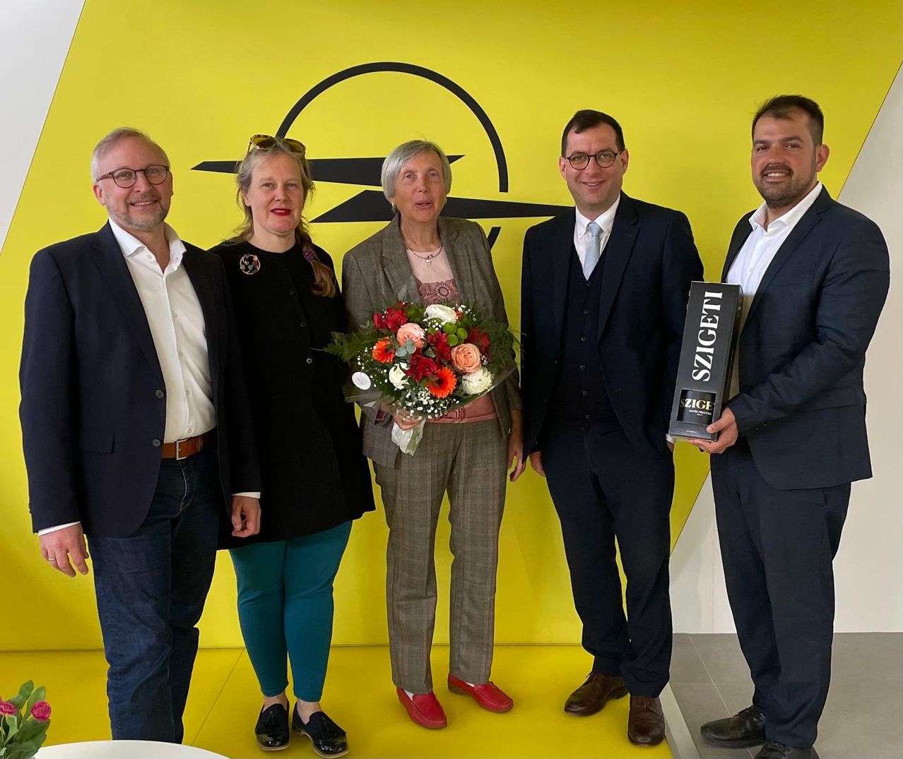 Fünf Personen stehen vor einem gelben Hintergrund und lächeln. Die Frau in der Mitte hält einen Blumenstrauß. Der Mann rechts hält eine Box mit der Aufschrift Zigeti.