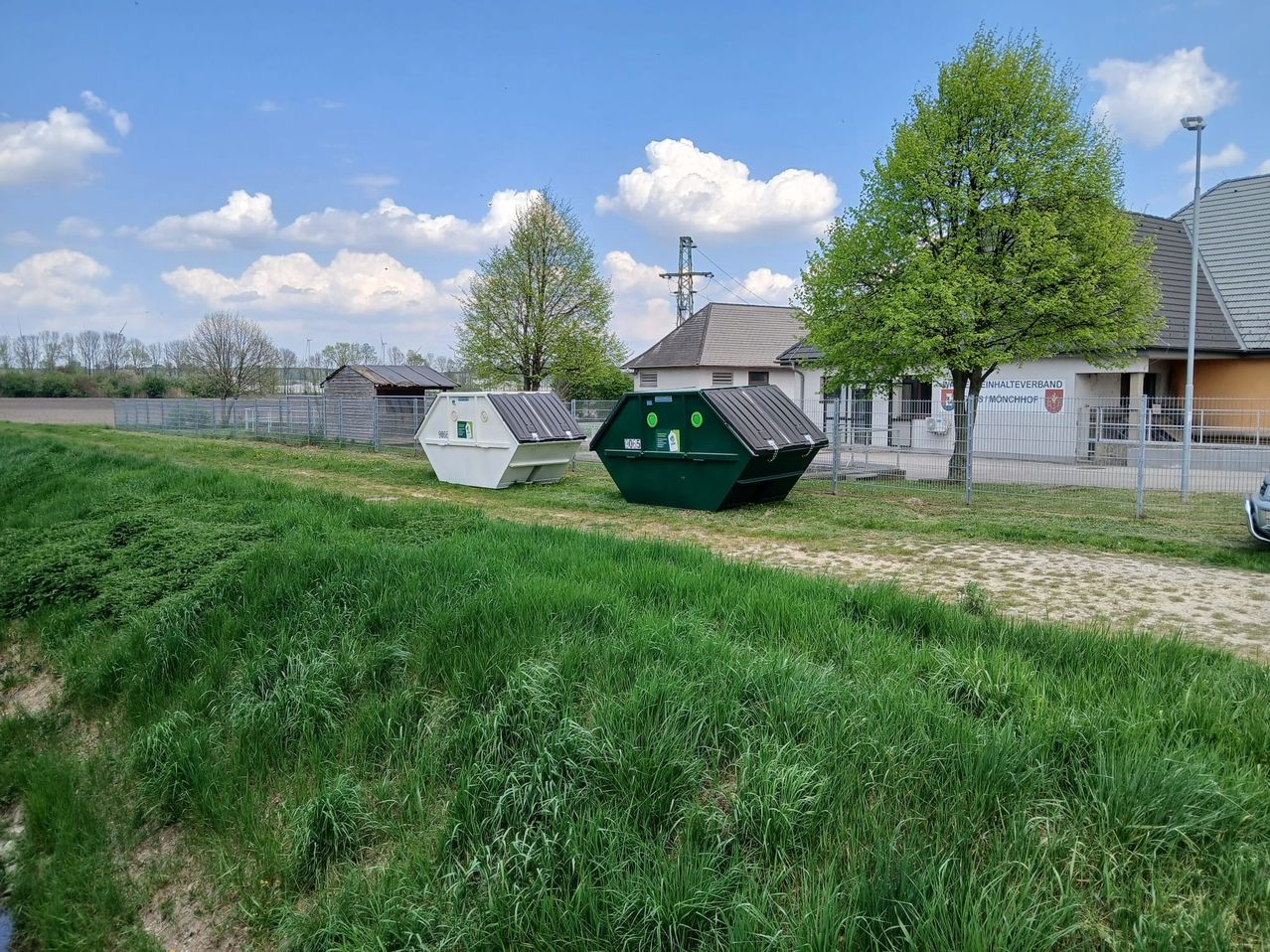 Zwei Müllcontainer stehen in einem grasbewachsenen Bereich mit einem Gebäude im Hintergrund. Bäume und ein Zaun umgeben das Gebiet, und der Himmel ist blau mit Wolken.