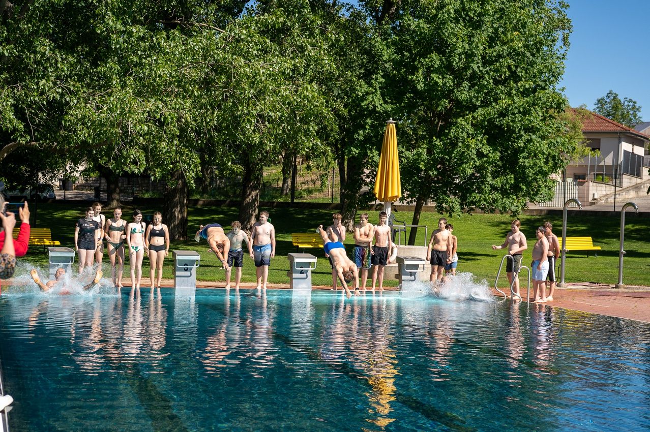 Eine Gruppe von Jungen und Mädchen steht am Rand eines Schwimmbeckens und bereitet sich auf den Sprung vor. Ein Junge springt bereits ins Wasser. Im Hintergrund befindet sich ein gelber Schirm und eine Bank.