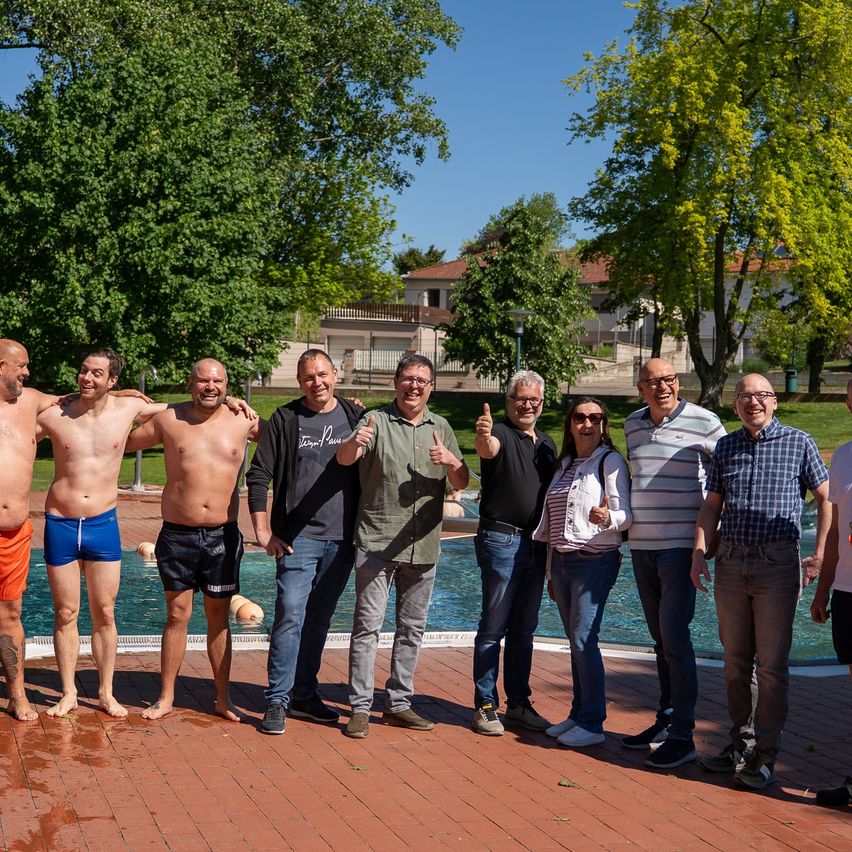 Eine Gruppe von Menschen steht in der Nähe eines Pools in einem Park, lächelt und posiert für ein Foto. Einige sind in Badeshorts, andere in Freizeitkleidung. Bäume und ein Gebäude sind im Hintergrund.