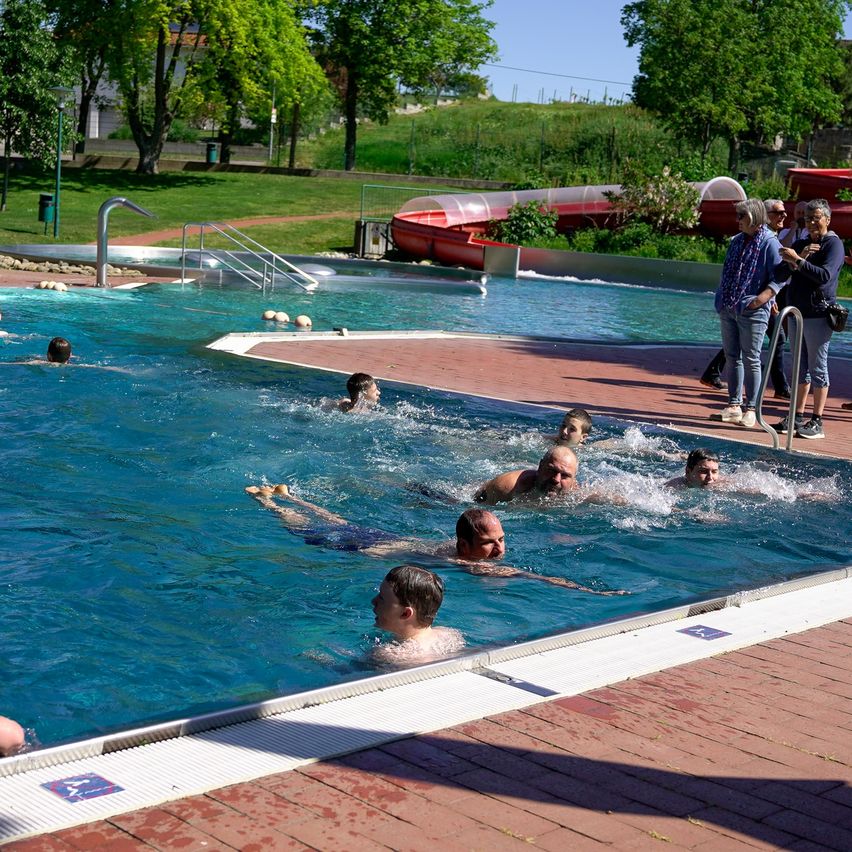 Eine Gruppe von Menschen schwimmt in einem Außenpool, eine Person liegt auf der Wasseroberfläche. Zuschauer stehen in der Nähe, einer hält eine Kamera. In der Nähe ist ein roter Wasserrutsche zu sehen.