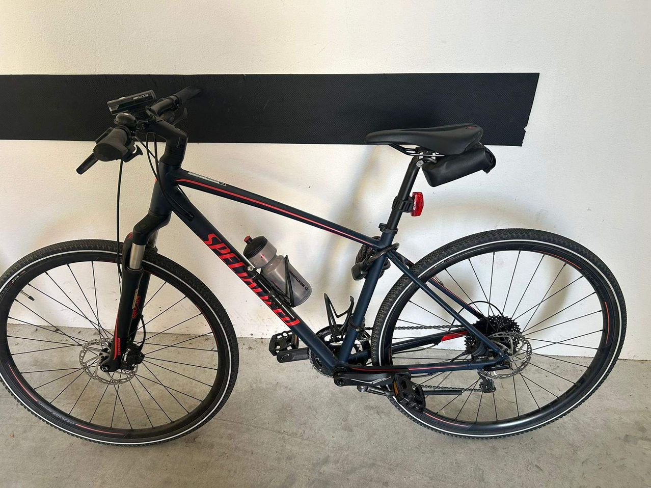 Ein schwarz-roter Mountainbike ist an einer weißen Wand mit einer schwarzen Matte geparkt. Eine Wasserflasche ist am Rahmen befestigt.