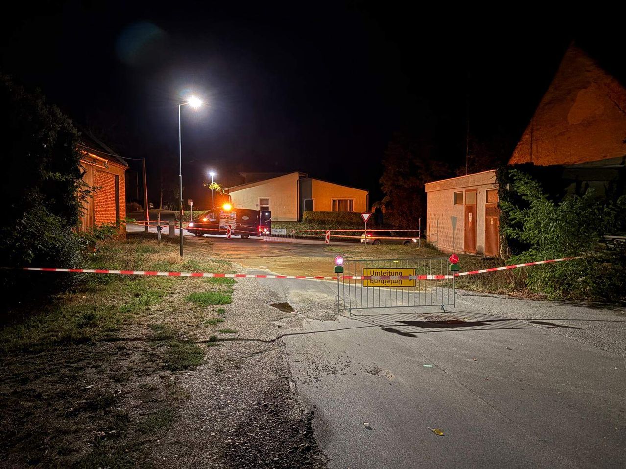 Eine Nachtszene mit einer gesperrten Straße, die durch eine Absperrung und blinkende Lichter blockiert ist. Ein Van ist in der Nähe geparkt und ein Haus ist im Hintergrund zu sehen.